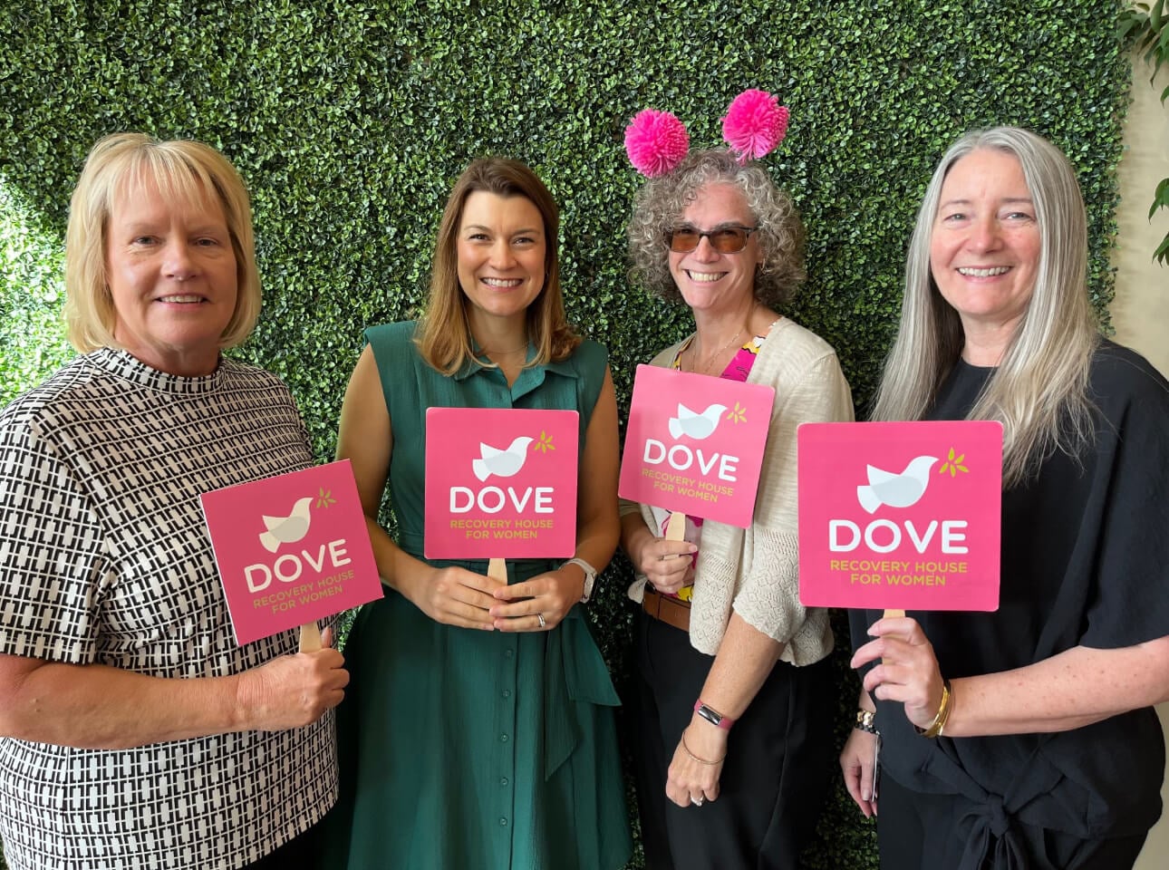 group of women standing in front of a green wall holding signs that say Dove Recovery House for Women