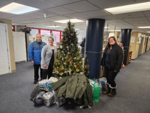 Three people standing near a Christmas tree with items for donation.